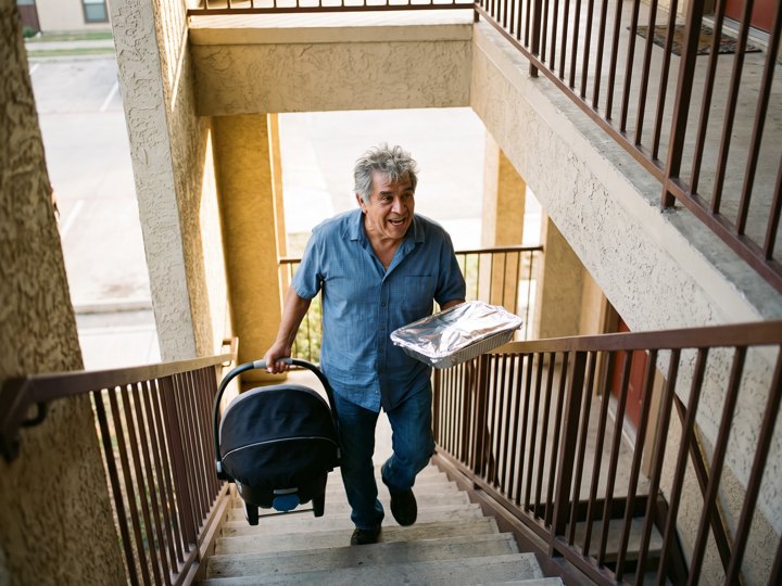 Un señor mayor subiendo una escalera exterior con una sillita de bebé y una olla tapada, sonriendo hacia atrás.